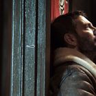 Ein Mann lehnt an einer Wand, hält ein Holzgewehr und blickt nachdenklich ins Licht, bekleidet mit einer Lederjacke.