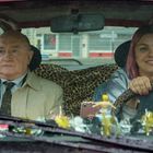 Ein älterer Mann und eine Frau mit rosa Haaren sitzen in einem Auto mit Leopardenmuster-Bezug, sie lächelt beim Fahren.