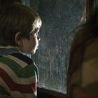 Kein Text sichtbar. Ein Kind im gestreiften Pullover blickt nachdenklich aus einem Fenster.