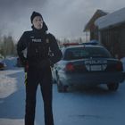 Polizist mit schwarzer Uniform vor einem Polizeiauto im Schnee, schneebedeckte Umgebung und wolkiger Himmel.