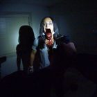 Eine Frau mit weit aufgerissenem Mund steht in einem dunklen Raum. Zwei Waffen sind auf sie gerichtet, sie trägt ein blutiges Shirt.