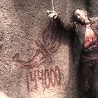 Die Wand zeigt rote Symbole und 1909. Eine Person hängt blutig an der Wand. Eine weitere Person sieht zu.