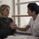 Zwei Frauen sitzen an einem Tisch. Eine trägt weiße Kleidung, hält die Hände der anderen und sieht sie tröstend an.