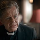 Ein Mann in Priesterkleidung sitzt drinnen, mit nachdenklichem Gesichtsausdruck, im Vordergrund. Im Hintergrund eine rote Lampe.