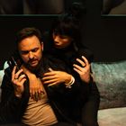 Ein Mann auf einem Sofa hält ein Telefon, während eine Frau ihn von hinten umarmt und ihn intensiv anschaut.