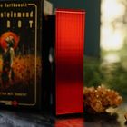 Text: "Steinmond Rot". Schwarze Verpackung mit Illustration. Roter Metallwürfel steht daneben auf dunkler Oberfläche.