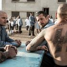 Text: "правда" Tätowierung. Zwei Männer in einem Gefängnishof an einem Tisch, umgeben von weiteren Insassen.