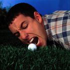 Ein Mann in kariertem Hemd liegt auf Gras und öffnet den Mund weit vor einem Golfball. Hintergrund ist blau.