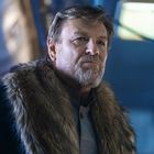 Ein Mann mit ernstem Ausdruck trägt einen Mantel mit Fellkragen in einer futuristischen Umgebung.