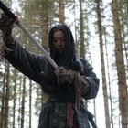 Eine Person in zerfetzter Kleidung hält ein Schwert in einem dichten Nadelwald. Die Stimmung wirkt ernst und konzentriert.