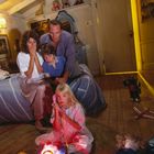 Ein Mädchen spricht in ein leuchtendes Spielzeugtelefon. Eine Familie schaut erstaunt zu. Spielzeug ist im Raum verteilt.