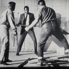 Drei Männer: zwei stehen mit Ketten und Rohren, einer im Anzug in der Mitte, konfrontativ. Szene in Schwarz-Weiß.