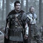 Zwei Soldaten in römischer Rüstung stehen in einem bewaldeten Gebiet, umgeben von gefallenen Kriegern.