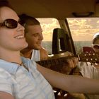 Eine Frau, ein Mann und eine weitere Frau sitzen fröhlich in einem Auto, die Frau rechts trägt eine Sonnenbrille.