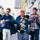 Vier junge Männer, einige mit Ballons, einer trägt ein Skateboard. Sie gehen auf einer städtischen Straße.