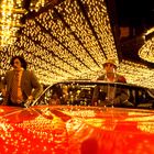 Keine Texte sichtbar. Drei Männer in auffälliger Kleidung stehen vor einem roten Auto. Helle Lichter umgeben die Szene.