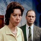 Zwei Personen: Eine Frau mit Perlenkette im Vordergrund und ein Mann im Anzug dahinter, vor blauem Hintergrund mit Pflanzenmotiv.