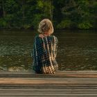 Eine Person in gemustertem Kleid sitzt auf einem hölzernen Steg am See, umgeben von Bäumen im Hintergrund.