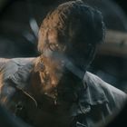 Ein Mann mit einer Maske wird durch ein zerbrochenes Glas betrachtet. Die Umgebung wirkt düster und rustikal.