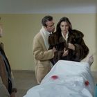 Eine Gruppe von Menschen steht um einen verdeckten Körper auf einer Bahre in einem kargen Raum.