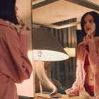 Kein Text zu sehen. Eine Frau in rosa Hemd trägt Lippenstift auf, steht vor einem Spiegel neben einer modernen Lampe.