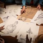 Skizzen von Figuren in historischer Kleidung auf einem Tisch, umgeben von Menschenhänden, die die Zeichnungen betrachten.