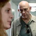 Zwei Personen in einem Flur, eine Frau im Vordergrund unscharf und ein Mann mit Sonnenbrille im Hemd schaut ernst.