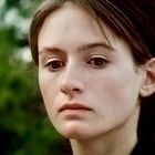 Eine Frau schaut ernst, mit natürlichem Hintergrund unscharf, vermutlich draußen.