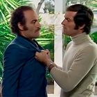 Zwei Männer stehen sich gegenüber; einer hält den anderen am Revers. Im Hintergrund sind tropische Pflanzen sichtbar.