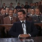 Ein Raum voller Männer in Anzügen, die in Reihen sitzen. Drei Männer sitzen im Vordergrund, wirken konzentriert.