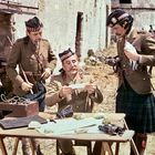 Drei Männer in Uniformen, einer liest ein Papier, ein anderer hält eine Taube. Ein Tisch mit Fernglas und Funkgerät.