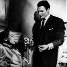 Ein Mann im Anzug reicht einer liegenden Frau ein Glas Wasser; sie trägt ein Kleid mit Volants, neben ihnen steht eine Lampe.