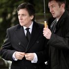 Zwei Männer in dunklen Mänteln, der linke hält sein Revers, der rechte hält einen Regenschirm über ihnen.