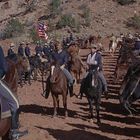 Keine Texte. Eine Gruppe von Reitern in blauen Uniformen, eine amerikanische Flagge wird getragen, Wüstenlandschaft im Hintergrund.