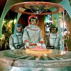 Drei Personen in silbernen Raumanzügen stehen um ein futuristisches Gerät mit leuchtendem Mittelpunkt.