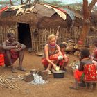 Ich kann den Text nicht erkennen. Eine Gruppe von Menschen sitzt auf dem Boden vor einer traditionellen Hütte aus Naturmaterialien.
