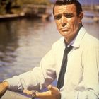 Krawatte, Hemd und Armbanduhr tragender Mann sitzt am Wasser, mit entspanntem Gesichtsausdruck und ausgestrecktem Arm.