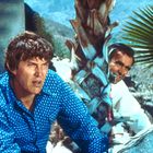 Zwei Männer mit ernsten Gesichtsausdrücken, im Hintergrund Palmen und blauer Himmel. Einer trägt ein blaues Hemd, der andere weiß.