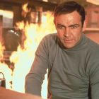 Ein Mann in einem grauen Rollkragenpullover, umgeben von Flammen, schaut ernst in die Kamera.