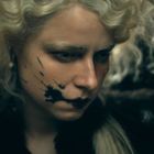 Keine sichtbaren Texte. Eine Person mit blonden, lockigen Haaren und schwarzer Gesichtsbemalung auf der Wange.