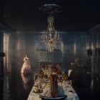 Ein elegant gedeckter Tisch in einem prunkvollen Raum. Eine Frau im Ballkleid steht; ein Kronleuchter hängt darüber.