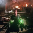 Ein Mann mit grünen Lichtern hebt sich aus einem brennenden, chaotischen Raum empor. Es wirkt actiongeladen und dramatisch.