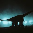 Silhouette eines Dinosauriers vor nebliger, bläulicher Lichterkulisse, Menschen spazieren im Vordergrund.