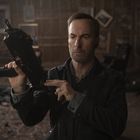 Ein Mann mittleren Alters mit ernstem Gesicht hält ein Gewehr in einem Raum voller Bilderrahmen.