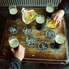Bierdeckel mit den Worten: PA, DDY, WAC, KERY. Gläser mit Getränken und geöffnete Chipstüten auf einem Holztisch. Hände greifen.