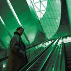 Eine Person in einem langen Mantel auf einer Rolltreppe in einem modernen Gebäude mit einer grünen Beleuchtung.