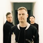 Drei Männer in einem engen, hellen Korridor. Der vordere trägt eine Jacke mit Kranich-Motiv.