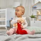 Ein Kleinkind sitzt auf einer Decke, hält ein Stofftier mit rotem Tuch, mit Krippe und Regal im Hintergrund.