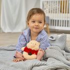 Kein Text vorhanden. Ein Kleinkind in blauer Kleidung sitzt auf einer grauen Decke, hält ein Kuscheltier und lächelt.