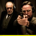 Zwei Männer mit Gehörschutz im Anzug, einer zielt mit Pistole nach vorne, ernste Miene, starker Kontrast.